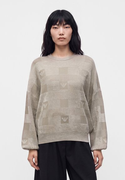 Femme aux longs cheveux noirs portant un pull beige ample à motif carré et un pantalon noir à jambes larges, debout devant un fond blanc uni.
