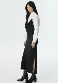 Vestido de satén negro con escote drapeado y abertura lateral, superpuesto a una camisa blanca. El modelo lleva botas negras brillantes hasta el tobillo.