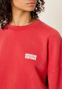 Sieviete ar cirtainiem matiem valkā sarkanu džemperi ar balto "AMERICAN VINTAGE" logotipu kreisajā krūtī, uz neitrāla fona.