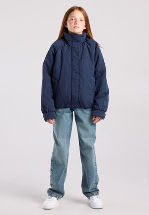 Enfant aux longs cheveux roux portant une veste bleu marine, un jean bleu clair et des baskets blanches, debout devant un fond clair uni.