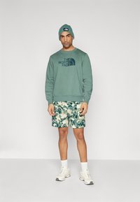 Hanorac verde cu logo-ul "The North Face", pantaloni scurți cu imprimeu camouflage în nuanțe de albastru și verde, adidași albi și o căciulă asortată.