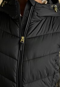 Czarna kamizelka puchowa o pikowanej fakturze, złotym suwaku i kapturem. Charakteryzuje się eleganckim designem i subtelnym brandingiem na metce obok zamka.