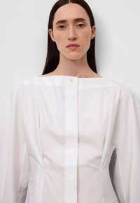 Witte blouse met een vierkante halslijn, lange mouwen en een enkele knoopsluiting aan de voren. Gemaakt van een gladde, gestructureerde stof.