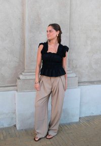 Γυναίκα στέκεται μπροστά σε πέτρινο τοίχο φορώντας μαύρο αμάνικο τοπ με βολάν, μπεζ παντελόνι με φαρδιά πατζάκια, σανδάλια και χρυσούς κρίκους στα αυτιά.