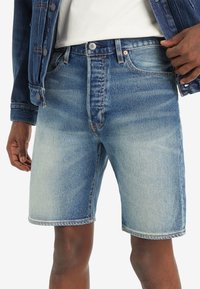 Shorts in denim azzurro chiaro con una finitura sbiadita, taglio dritto, design a cinque tasche e dettagli di cuciture visibili. Indossati con una giacca di denim.