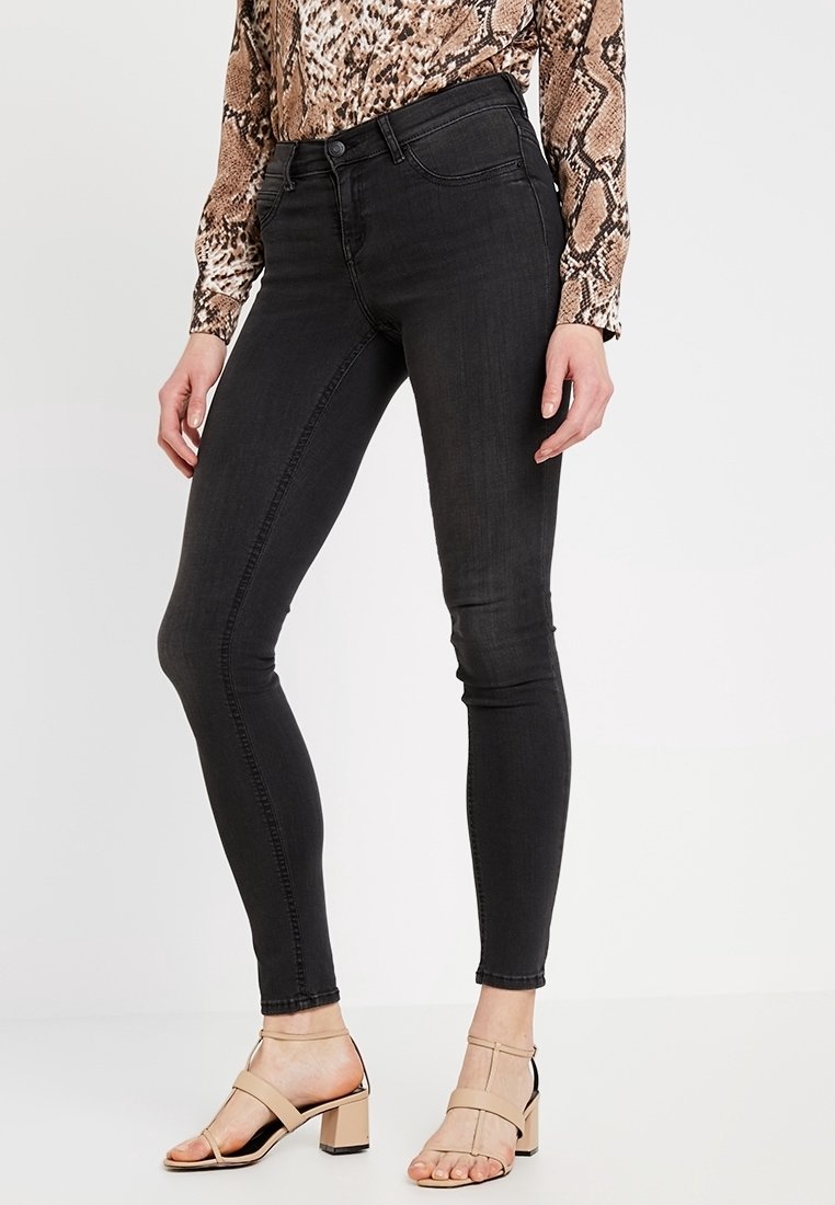 Jean skinny noir avec une texture lisse, taille haute et design minimaliste. Porté avec des sandales à talons blocs nudes.