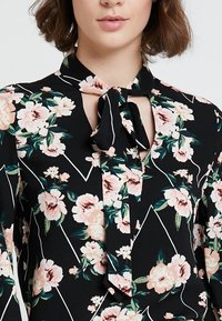 Blusa nera con stampa floreale che presenta rose rosa e bianche. Dettaglio con fiocco al collo e accenti a linee geometriche bianche.