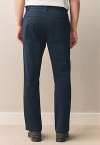 Pantalon bleu marine en coton, présentant une coupe droite, deux poches arrière et une texture lisse. Porté avec des chaussures marron.