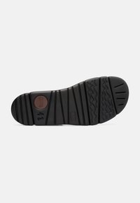 Sola in gomma nera con pattern testurizzati, scanalature segmentate e un logo circolare marrone. Presenta varie linee di presa e una punta arrotondata.