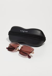 Occhiali da sole quadrati con lenti marroni, dettagli in oro e punte bordeaux, accompagnati da un astuccio nero con zip contrassegnato con "vogue".