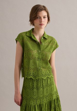 Junge Frau trägt grünes Häkel-Button-up-Top und passenden Rock, steht vor neutralem einfarbigem Hintergrund.
