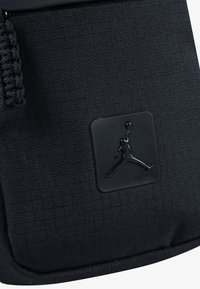 Black textured fabric bag featuring a raised black Jumpman logo patch and a braided black cord accent. Compact design, minimalistic style.