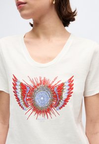 Γυναίκα που φοράει λευκό T-shirt με V λαιμόκοψη και λαμπερό κόκκινο, μπλε και χρυσό κυκλικό σχέδιο με φτερά στο στήθος, με εμφανές χρυσό κρίκο σκουλαρίκι.