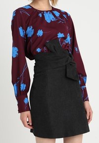 Blouse bordeaux à imprimé floral bleu, manches longues ; associée à une jupe portefeuille noire avec un détail de nœud à l'avant.