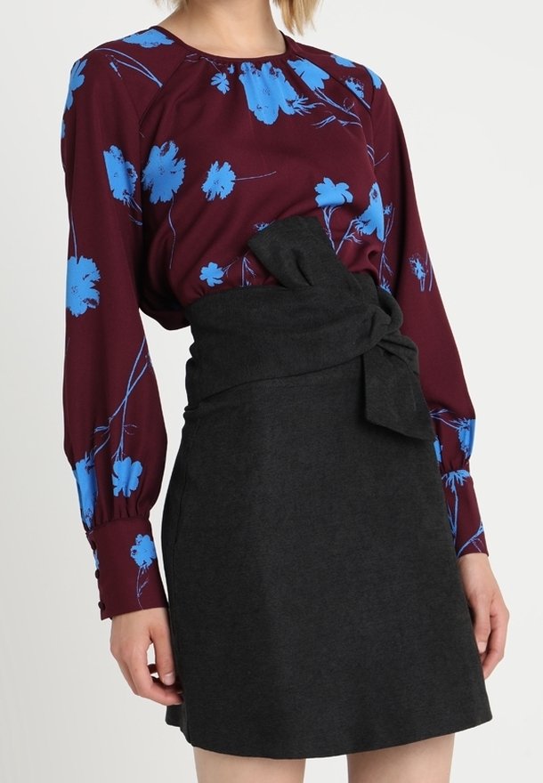 Blouse bordeaux à imprimé floral bleu, manches longues ; associée à une jupe portefeuille noire avec un détail de nœud à l'avant.