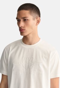 Valkoinen puuvillateeppari, jossa on pyöreä kaula-aukko. Edessä on kohokuvioitu GANT-logo ja "AMERICAN SPORTSWEAR" -teksti.