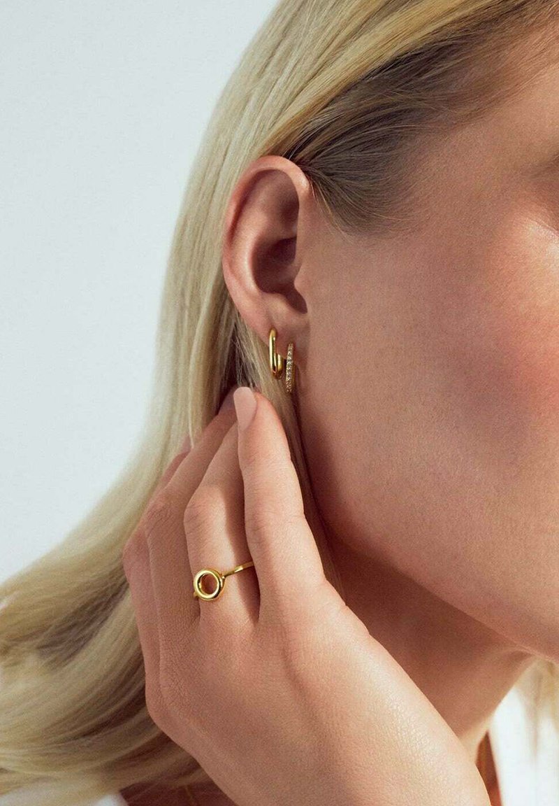 Blond kvinna som rör vid örat med handen och bär guld-hoopörhängen samt en guldring med cirkulär design på fingret.