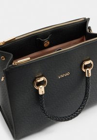 Sort struktureret håndtaske med en tekstureret finish, guldfarvet hardware, flettede håndtag og en rosa indvendig foring. Logoet er trykt på forsiden.