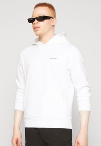 Sweat-shirt à capuche blanche à coupe ajustée, avec logo Calvin Klein en gris. Fabriqué en tissu doux, avec des manches longues et des poignets côtelés.