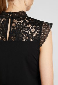 Zwart top met ingewikkelde kanten details op de schouders en rug, met een knoopsluiting in de nek en geschulpte kanten randen.