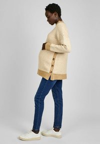 Maglione a motivo chevron di un beige chiaro e crema con bottoni laterali e orlo a costine, abbinato a jeans blu scuro e sneaker bianche.