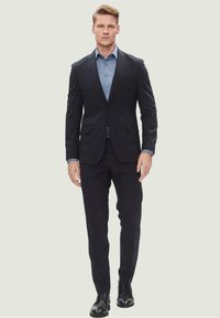 Abito navy su misura con rever a punta e chiusura a un bottone. Indossato sopra una camicia azzurro chiaro con bottoni, abbinato a scarpe nere.