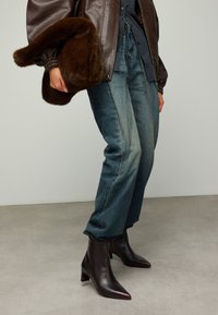 Bruine leren pilotenjack, blauw geruit overhemd, vervaagde blauwe spijkerbroek en donkerbruine enkellaarsjes met spits toelopende neuzen en blokhakken.