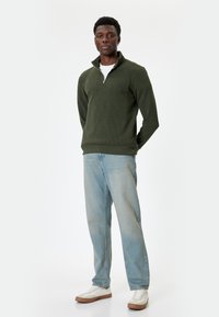 Pullover verde con zip e texture a coste, abbinato a jeans azzurri a gamba dritta e sneakers bianche con dettagli marroni.