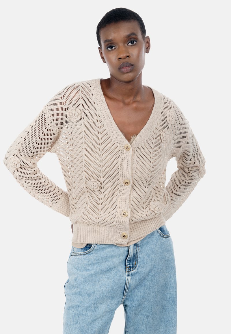 Beige strikket cardigan med v-hals, knaplukning, åben vævemønster og blomsterdetaljer. Kombineret med lyse denimjeans.