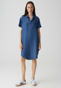 Denimhemdkleid in Mittelblau, mit Knopfkragen, kurzen Ärmeln, einer Tasche auf der linken Brust und abgerundetem Saum. Getragen mit weißen Sneakers.