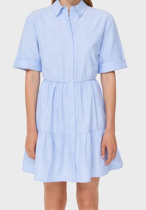 Mujer que lleva un vestido azul claro de manga corta, con cuello, abotonado, con cintura fruncida y falda de volantes.