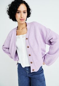 Cardigan leggero viola chiaro con texture a coste, chiusura a bottoni, taglio oversize, indossato sopra un top bianco con bordo in pizzo e abbinato a jeans blu.
