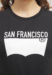 Svart bomullströja med en vit grafik som visar texten "SAN FRANCISCO" och en stiliserad logotyp under, rund halsringning och korta ärmar.