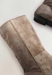 Bottes mi-mollet en daim taupe avec des détails de couture le long de la tige et des talons bas en caoutchouc noir sur un fond clair.