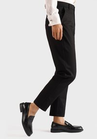 Pantaloni neri con una vestibilità su misura, trama del tessuto visibile; abbinati a mocassini neri lucidi con una fascia decorativa.