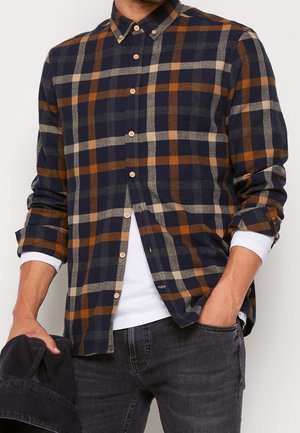 Camisa de franela a cuadros azul y naranja con cuello de botón, que cuenta con botones de madera y mangas arremangadas, llevada sobre una camisa blanca.
