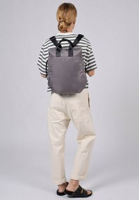 Mochila de tela acolchada gris con correas negras, asa superior y cierre de cordón, vista desde la parte trasera en una persona que lleva una camisa a rayas y pantalones crema.