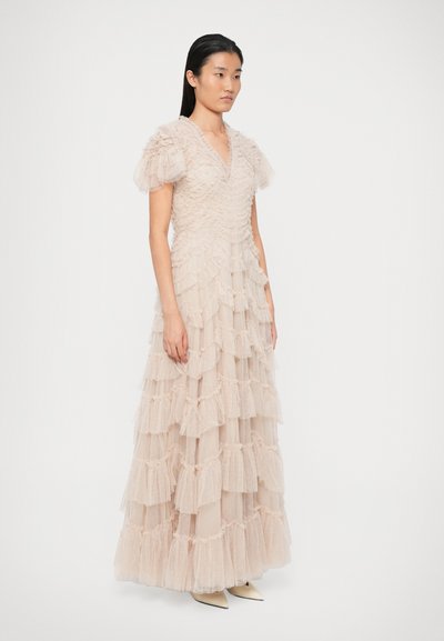 Γυναίκα που φοράει ένα πατωματαίο, ανοιχτό μπεζ τούλινο φόρεμα με επίπεδες ρίγες και κοντά φουσκωτά μανίκια, στέκεται μπροστά σε έναν απλό λευκό φόντο.