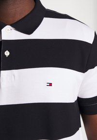 Polo shirt rayé noir et blanc avec un col, comportant deux boutons et un petit logo brodé en rouge, blanc et bleu sur la poitrine.