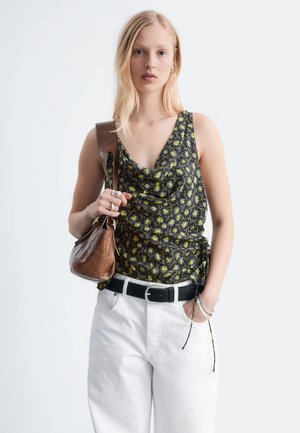 Mladá žena v čiernom kvetovanom tielku bez rukávov, bielych nohaviciach, čiernom opasku, držiaca hnedú koženú kabelku cez plece.
