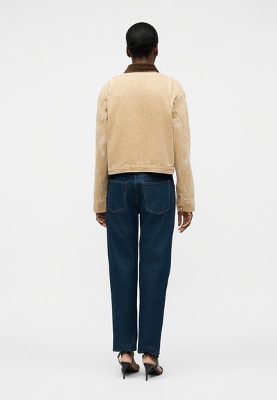 Άτομο με σκουρόχρωμο δέρμα και μαλλιά δεμένα πίσω, φοράει μπεζ μπουφάν, σκούρο μπλε τζιν και μαύρα σανδάλια με τακούνι, στέκεται με την πλάτη προς τα εμάς.