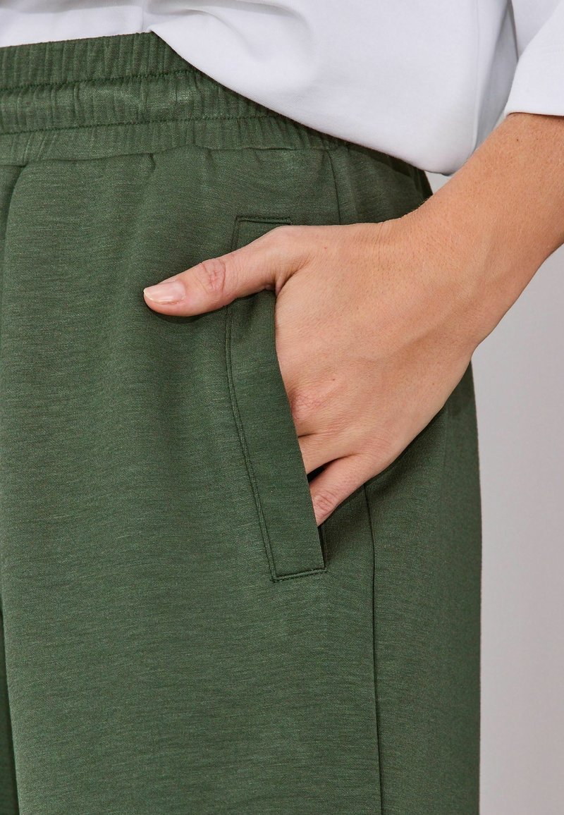 Ruka so svetlým odtieňom pokožky odpočívajúca v priehradke zelených elastických teplákov s opaskom, oblečená v bielom tričku s dlhým rukávom.