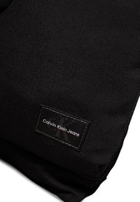 Svart stoffveske med en teksturert overflate og en synlig etikett med teksten "Calvin Klein Jeans" med en sydd kant. Utsmykket med en logo.