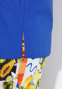 Top in cotone blu con una fessura laterale, sopra pantaloni corti colorati a fantasia con disegni di frutta su uno sfondo chiaro.