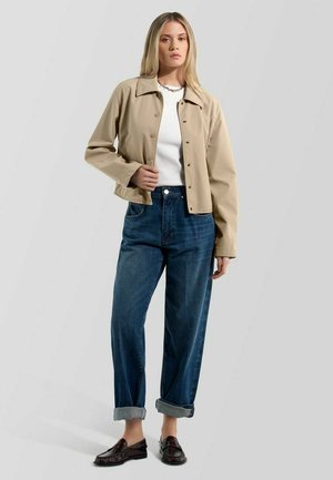 Frau trägt beige Jacke mit Knöpfen, weißes Oberteil, hochgekrempelte blaue Jeans und dunkelbraune Loafers, steht vor einfarbigem Hintergrund.