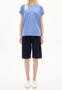 Jasnoniebieski t-shirt z krótkim rękawem ze zrolowanymi mankietami w połączeniu z ciemnymi, dżinsowymi szortami sięgającymi do kolan, noszony z białymi trampkami.
