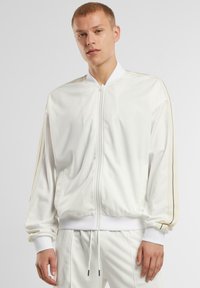 Giacca bomber bianca in tessuto liscio, con zip frontale, polsini e orlo a costine. Presenta righe beige a contrasto sulle maniche e sulle tasche.