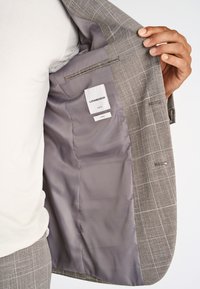 Main tenant une veste de costume beige à carreaux ouverte, montrant une doublure intérieure grise lisse et une étiquette indiquant "Lindbergh Slim Fit Stretch".