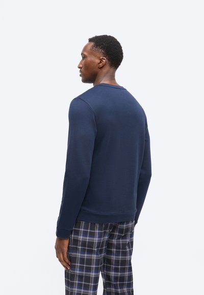 Άντρας που κοιτά προς τα αριστερά, φορώντας μπλε σκούρο φούτερ και καρό παντελόνι σε μαύρο, μπλε και άσπρο, σε απλό λευκό φόντο.