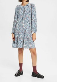 Blommig klänning med lång ärm, rund halsringning, flikig fåll, främst ljusblå med rosa och gröna inslag. Bärs med svarta skor och bourgognefärgade strumpor.